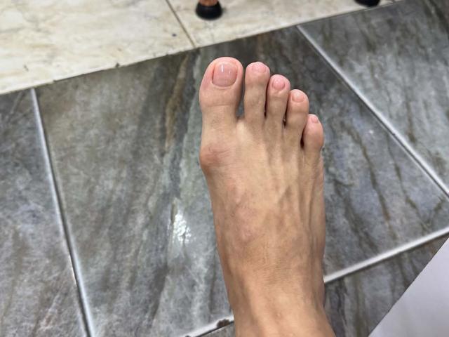 Il piede di JoseRumBerotop, che mostra un solo alluce con un'unghia leggermente rosa, pulita e curata, poggiato su piastrelle di marmo grigio con sottili venature bianche..