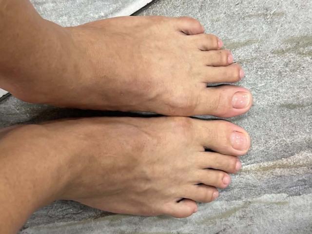 Os pés de JoseRumBerotop, mostrando dois dedos em cada pé com unhas levemente rosadas, repousam sobre uma superfície cinza texturizada. A pele é de cor marrom clara e os dedos estão limpos..