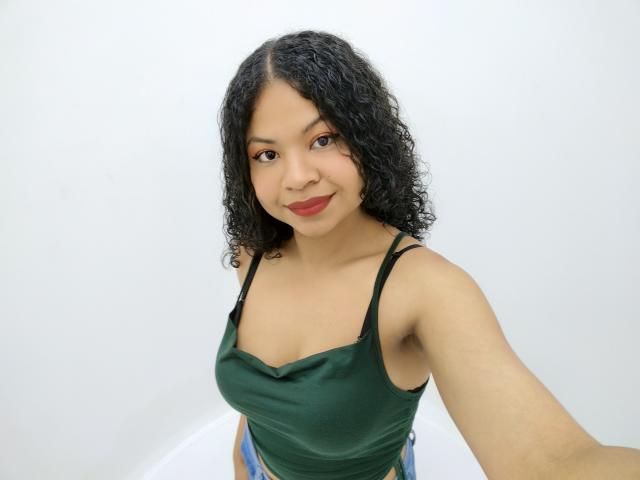 Nhicol, met krullend zwart haar, draagt een groene tanktop, toont kleine borsten, glimlacht met rode lippenstift, houdt een telefoon vast voor een selfie, op een witte achtergrond..