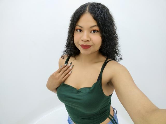 Nhicol, met krullend zwart haar, draagt een groene tanktop, waarbij haar borsten zichtbaar zijn, met een hand op haar borst, blauwe geruite broek, rode lipstick, lachend, lichte huid, selfie..