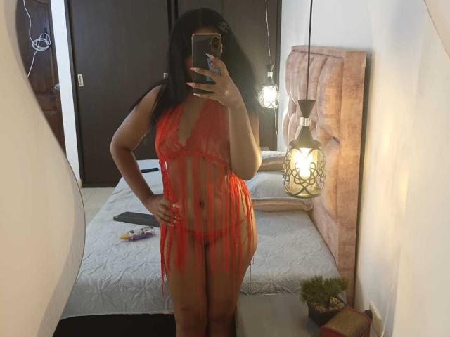 MiaStClaire with medium tits and a small ass, wearing a sheer red lingerie dress, taking a mirror selfie, holding a phone, in a dimly lit bedroom.