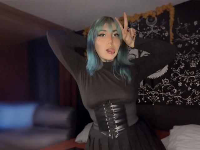 AliceCalloway with blue-green hair, black dress, and black corset, making a peace sign with her right hand, has a cute face and a small nose ring.