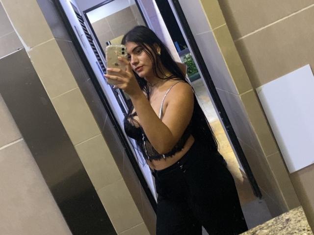 Alara, con el pelo largo y oscuro, vestida con un sujetador de encaje negro y pantalones negros de cintura alta, se está tomando un selfie frente al espejo con un teléfono inteligente, mostrando sus pechos de tamaño medio y una sonrisa sutil..