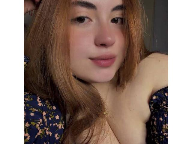 VicluX with light brown hair, slightly parted lips, and a subtle smile, wearing a floral dress, showing her tits, wearing a gold necklace, in a selfie.
