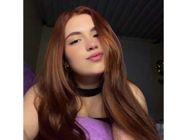 VicluX has long, red hair, a black choker, and is smiling slightly with pink lips. She's wearing a purple top, and the shot is a low-angle selfie.