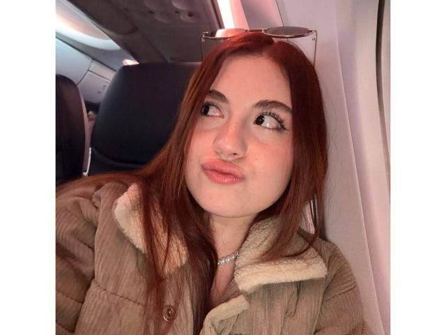 VicluX with long red hair, wearing a puffy beige coat with a fur collar, sitting in an airplane, looking up with pouty pink lips, and glasses on her head.