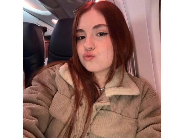 VicluX has red hair, pale skin, and is pouting her lips. She's wearing a beige, quilted, fur-lined jacket with a buttoned front. She's in an airplane.