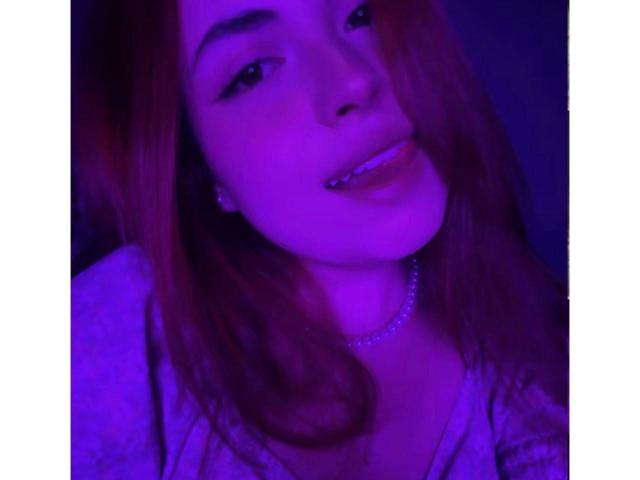 VicluX, avec un visage aux reflets rosés et violets, des cheveux bruns, un sourire discret, des dents visibles avec un seul piercing, un collier de perles et un haut blanc, dans un selfie aux couleurs bleutées et à faible luminosité..