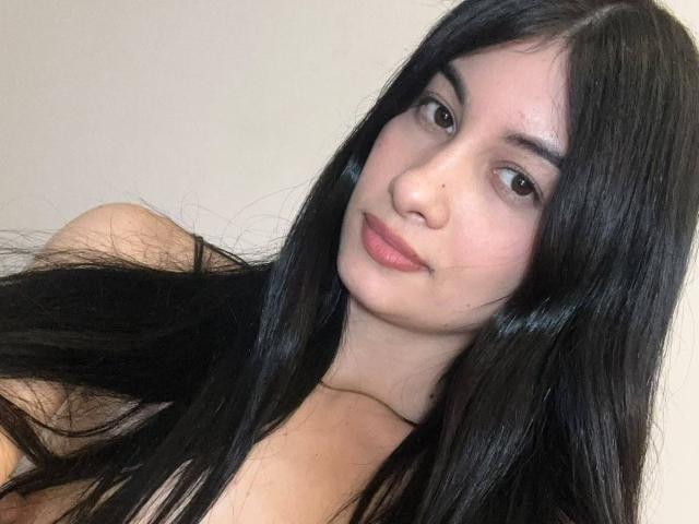 DevineSimone has long black hair, brown eyes, and a slight smile. She's looking at the camera with a relaxed, sexy, and attractive face, showing her bare shoulders.