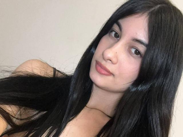 DevineSimone with long black hair, brown eyes, and pink lips, looking slightly to the side, showing a calm, subtle smile. Her bare shoulders are visible.
