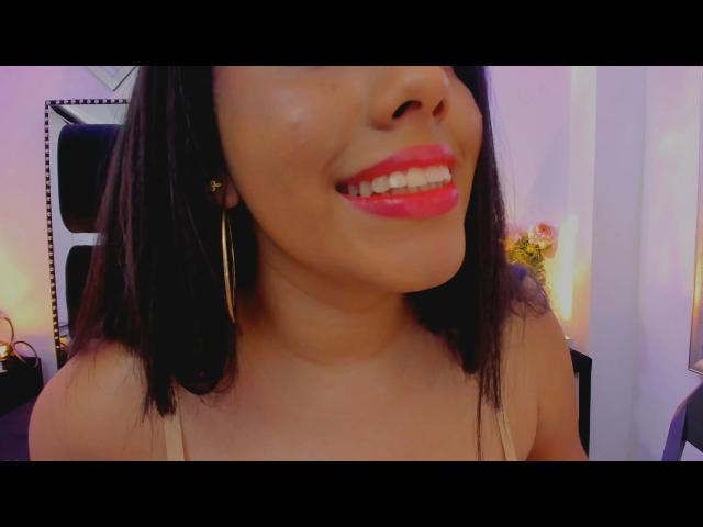 SiennaTaylor has shiny black hair, red lipstick, and is smiling with visible teeth. She's wearing gold hoop earrings and a tan spaghetti strap top.