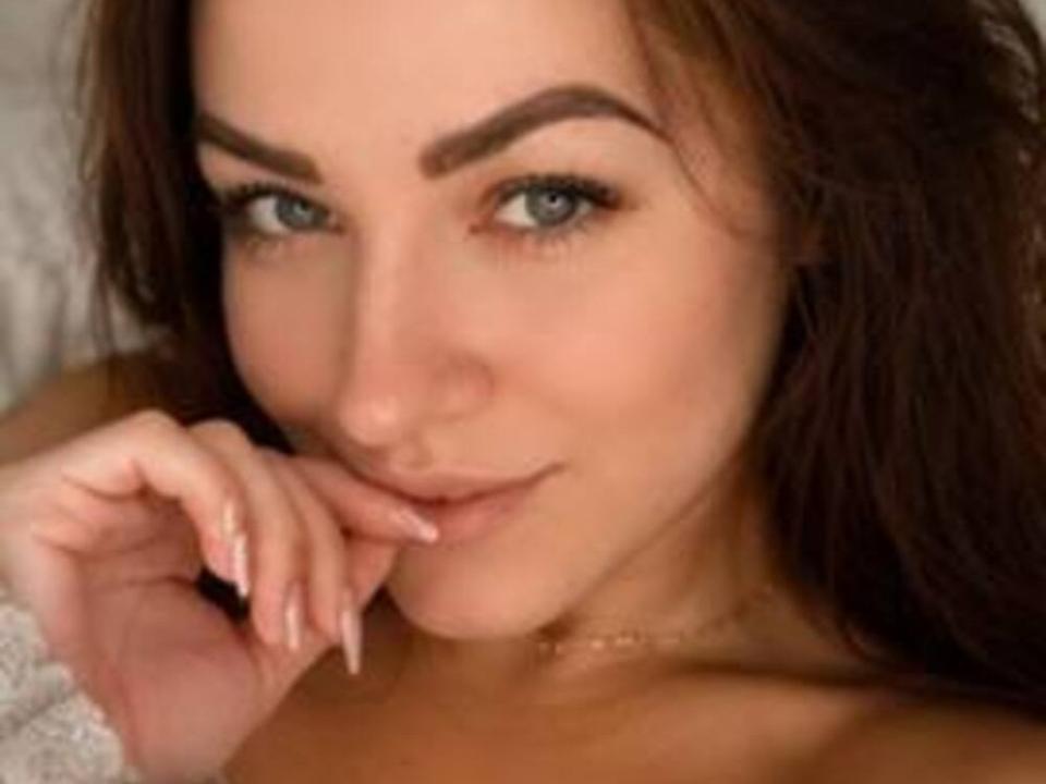BonnyDanielas has blue eyes, dark brown hair, and is smiling with her finger touching her lip. She has a cute nose and is wearing a gold necklace.