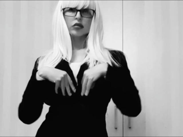 NattyNat-hot with pale blonde hair, black glasses, and a black zip-up sweater, hands near chest, black-and-white photo, showing tits, slightly open lips, wooden background.
