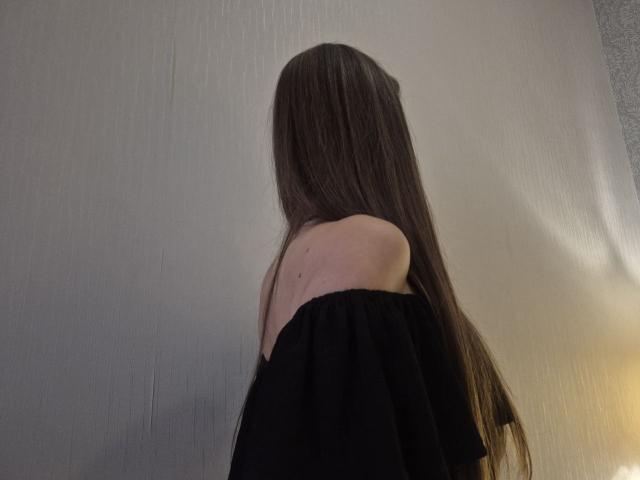 TenerliRize with long brown hair, off-shoulder black top, showing bare shoulders and upper back, facing away, in a dimly lit room.