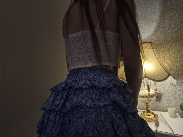 TenerliRize with dark hair, wearing a sheer purple top and a blue, ruffled, lace dress, standing in front of a lit, golden lamp. TenerliRize's ass is prominent.