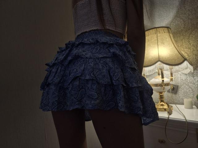 TenerliRize's blue, ruffled, lacey skirt is up, showing her bare ass. She's wearing a grey, textured top, and a vintage-style lamp is on the right.