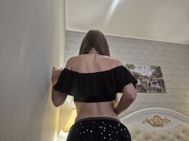 TenerliRize with back to camera, black off-shoulder top, showing her bare back and small tattoo, black polka dot pants, standing against a wall, long brown hair.
