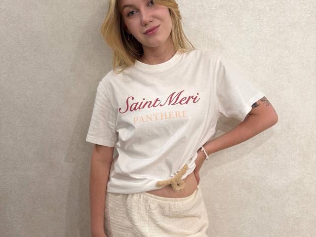 RudyPosada, blonde, portant un t-shirt blanc avec l'inscription "Saint Meri Pantherelle" en rouge, noué à la taille, un pantalon blanc duveteux, un petit tatouage sur le bras droit, un léger sourire..