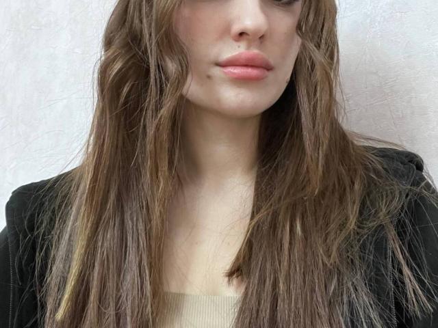 Lennie Fardo, avec ses longs cheveux bruns et indisciplinés, porte une veste noire par-dessus un haut beige. Elle a des lèvres rosées et une petite ecchymose discrète sous son œil droit..