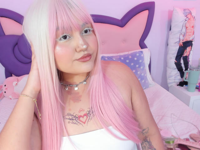MarlaBriard with pink and white long hair, wearing a white strapless top, showing tattoos, nose ring, and necklace, sitting on a pink and purple bed. Boobs visible.