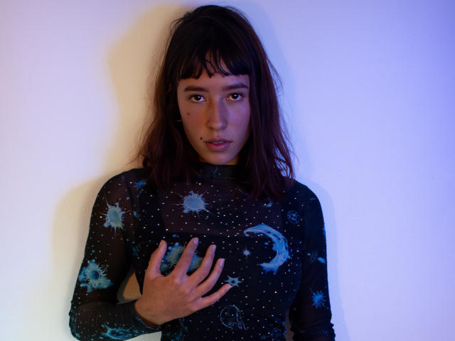 JuliethCourtouis with dark brown hair, wearing a black, starry, moon-patterned, sheer top, standing against a white wall, hand on chest, looking directly at the camera.