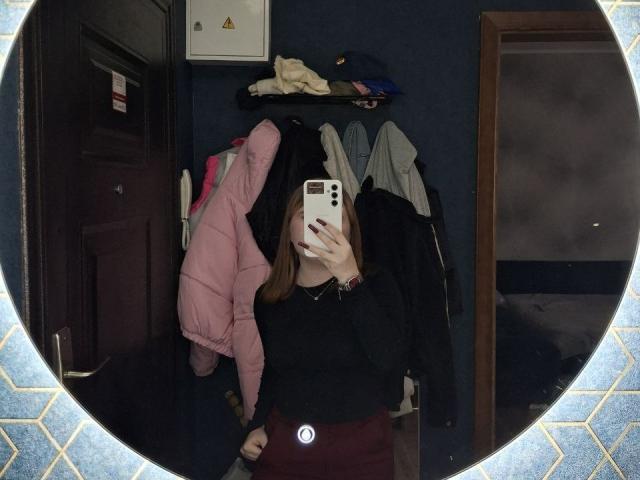 AlysiaFillingham si scatta un selfie allo specchio, coprendo il viso con un telefono bianco, indossando una maglietta nera e una giacca rosa, in piedi in una stanza buia piena di vestiti..