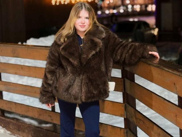 Alysia Fillingham, avec de longs cheveux blonds, portant un manteau en fourrure marron, un pantalon bleu, appuyée contre une clôture en bois, nuit, debout, texture épaisse du manteau, expression détendue..