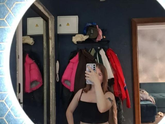 AlysiaFillingham tirando uma selfie em um espelho, mostrando seus seios em uma blusa preta com alças finas, com um celular cobrindo seu rosto, e roupas penduradas em uma parede escura..
