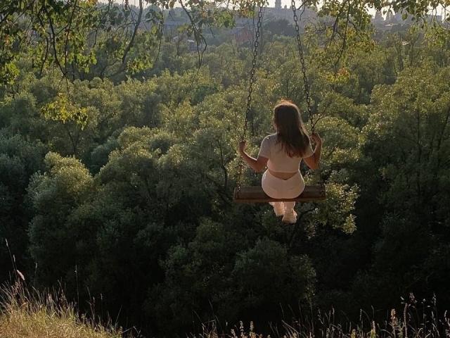 Alysia Fillingham è seduta su un'altalena di legno con catene di metallo, con la schiena rivolta verso la telecamera, indossa un vestito bianco e il suo sedere è leggermente sollevato, in una foresta verde illuminata dal sole..