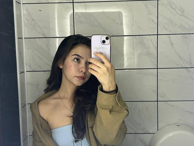 AnneKofron with long brown hair, light skin, wearing a green zip-up jacket and blue strap top, taking a mirror selfie with a white phone in a tiled bathroom.