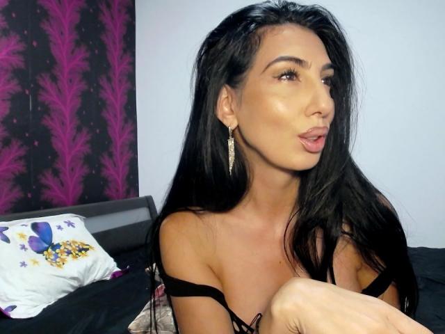 BrunetteBabe69 with long black hair, wearing a black strap top, pouty lips, and gold earrings, has a sexy tan skin tone and is sitting on a bed with a pillow.