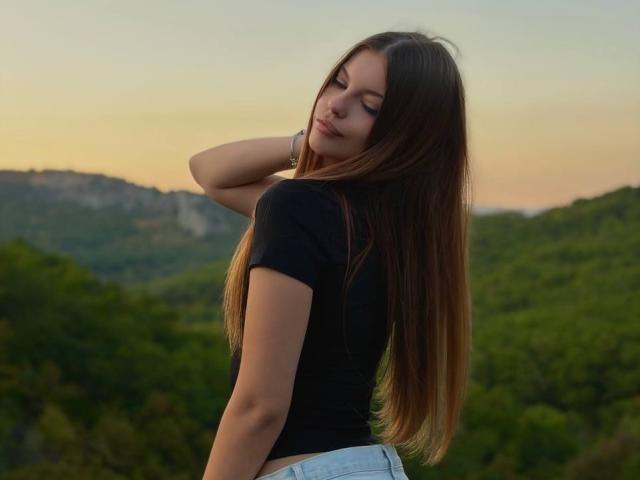 BethanySandhoff with long brown hair, black top, and blue jeans, looking back with a slight smile, hand in hair, standing in a green forest at sunset.