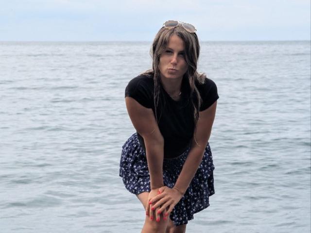 AuroraStar with wet brown hair, black t-shirt, and blue floral skirt, leaning forward, hands on knees, red nails, sunglasses on head, looking serious, standing in shallow ocean.