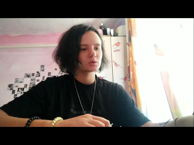 TreenaVisosky with messy black hair, wearing a black shirt, necklace, and bracelet, sitting near a bright window, looking slightly bored or distracted.