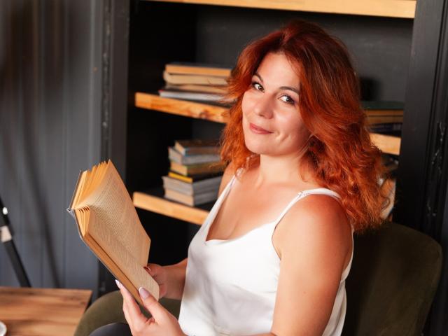 ZheniaNikrasova mit roten Haaren, einem weißen Tanktop, die ein offenes Buch hält, leicht lächelnd, sitzend auf einem grünen Stuhl, die in die Kamera schaut und im Hintergrund ihr Gesäß zeigt..