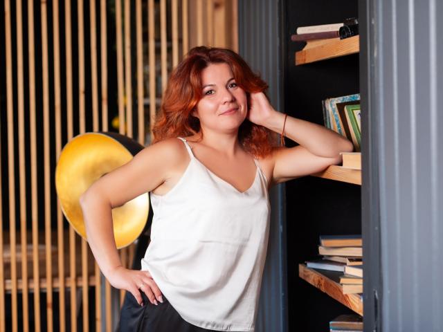 ZheniaNikrasova mit roten, lockigen Haaren, weißem Tanktop, die an einem Bücherregal lehnt, lächelnd, Hand am Wangen, schwarze Hose, Bücher, Holzregale, goldene Lampe, entspannt, süß, lässig..