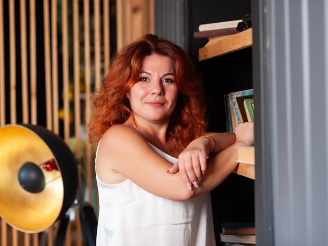 Zhenia Nikrasova mit roten, lockigen Haaren, einem weißen Top ohne Ärmel, mit verschränkten Armen, lächelnd, steht neben einem Bücherregal, eine goldene Stehlampe ist links, im Hintergrund sind Holzlatten zu sehen..