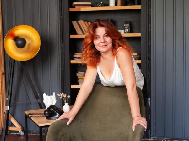 Zhenia Nikrasova mit roten, lockigen Haaren, einem weißen Tanktop und einem olivgrünen Rock, die an einem Stuhl lehnt, lächelnd, mit leicht angehobenem Gesäß, in einem gemütlichen, beleuchteten Raum..