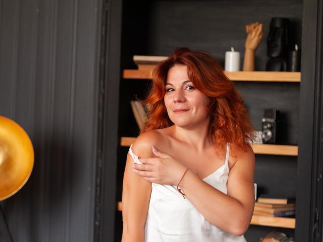 Zhenia Nikrasova mit roten Haaren, die ein weißes Tanktop trägt, bedeckt ihre kleinen Brüste mit einer Hand, lächelt leicht und steht vor einem schwarzen Regal..