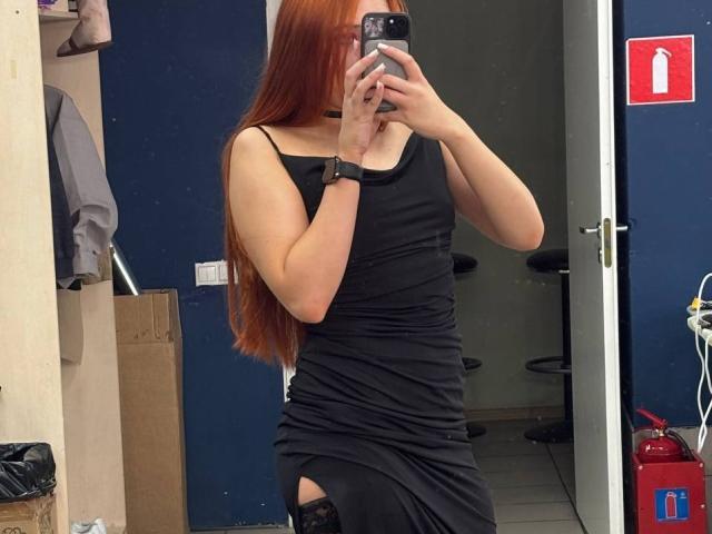 PeiSakasegawa with long red hair, wearing a black dress, showing a thigh cutout, black choker, and wristwatch, taking a mirror selfie in a room.