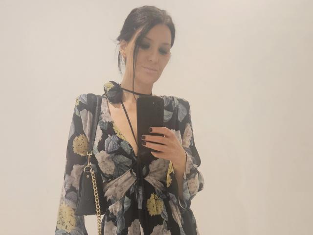 JoliDanielle, con i capelli scuri raccolti in uno chignon, indossa un abito nero con fiori e un profondo scollo a V, tiene in mano un telefono nero, ha lo smalto nero sulle unghie e porta una borsa a catena dorata..