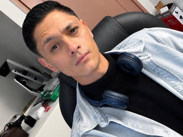 TylerCa, short black hair, brown eyes, light skin, wearing a black turtleneck, light blue denim jacket, blue headphones around neck, black leather chair, slightly confused expression.