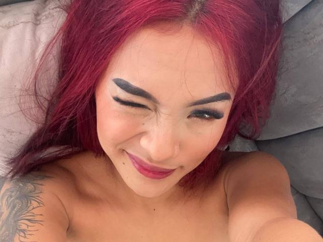 SashaHans with red hair, winking, thick black eyebrows, tattoo on right shoulder, pink lipstick, smiling, topless, visible collarbone, lying on gray pillows.