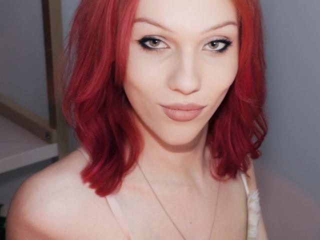 KiranaHayes with red hair, pale skin, and black eyeliner, wearing a white top, has a neutral expression and small, slightly upturned nose, showing her tits and a silver necklace.