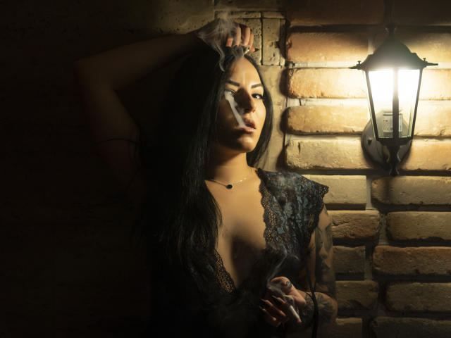JustIzabel, com longos cabelos pretos, vestindo uma blusa de renda, fumando, segurando um cigarro, braço levantado, expondo seus pequenos seios, colar, luminária de parede, iluminação escura e melancólica, parede de tijolos..