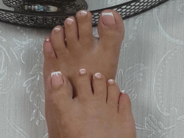 Os pés da JustIzabel, com unhas dos pés pintadas no estilo francês, mostrando esmalte rosa e pontas brancas, posicionados sobre uma superfície de madeira, com uma cesta de metal ao fundo..