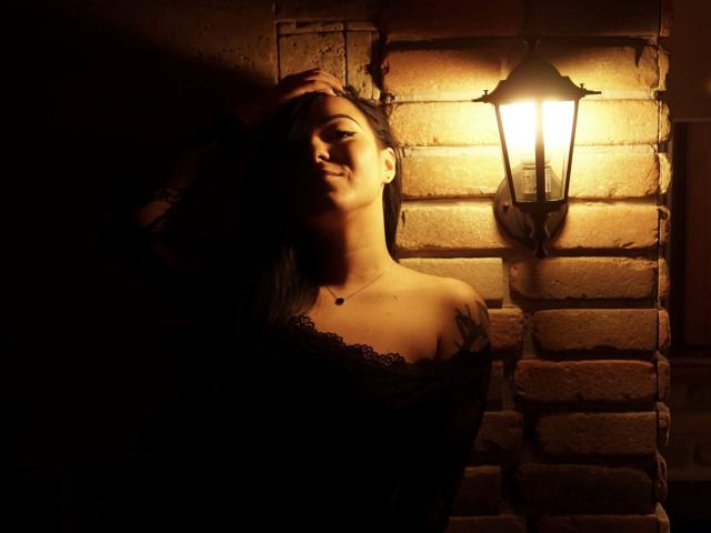 JustIzabel, con i capelli scuri, in piedi nell'ombra, con una mano sulla testa, indossa una camicetta di pizzo nero. La luce di una lanterna illumina il suo seno e il tatuaggio sulla spalla destra. Illuminazione calda e soffusa..