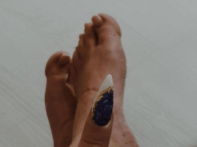 O dedo sujo do pé da JustIzabel, com uma escova de dentes branca com cerdas azuis e douradas, segurado pelo dedo dela, em um chão branco..