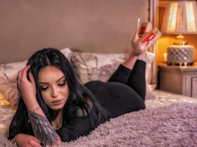 JustIzabel lies on a bed, black hair, tattooed arm, wearing black pants, beige heels with red soles, looking down, ass up, hand in hair, purple blanket, warm lighting.