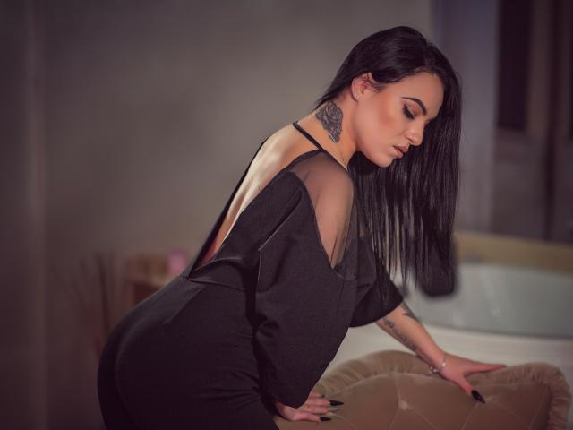 JustIzabel con i capelli neri, vestito nero senza schienale, tatuaggio visibile sul collo, appoggiata a un divano beige, che mostra il sedere, unghie nere, profilo laterale, illuminazione soffusa..